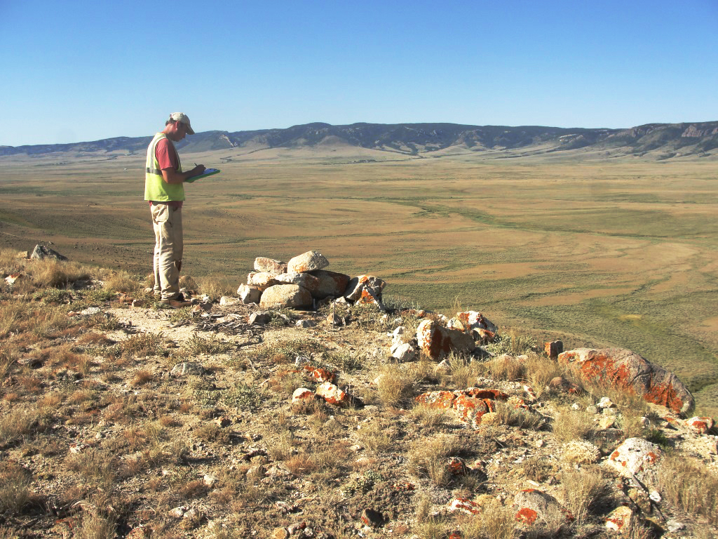 Crewmember standing at edge of steep slope with open field and mountains in distance.
