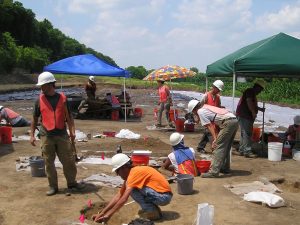 CRA crew conducting fieldwork in open excavated area, including shade tents and an umbrella.