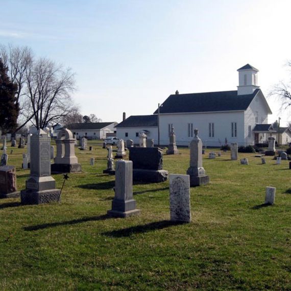 Overview of historic cemetery, featuring several obelisk gravestones with church in background.