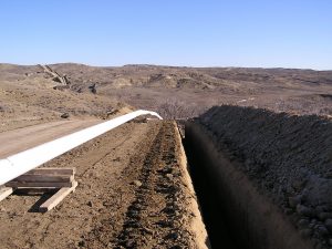 Pipeline trench next to dirt road, with mountainous terrain in background.