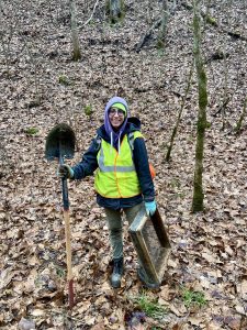 Millie standing in leaf-covered area of woods, holding a shovel and a soil screen.