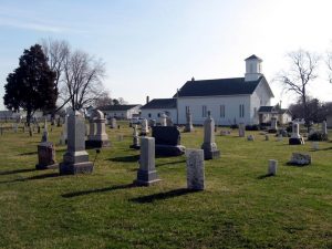 Overview of historic cemetery, featuring several obelisk gravestones with church in background.