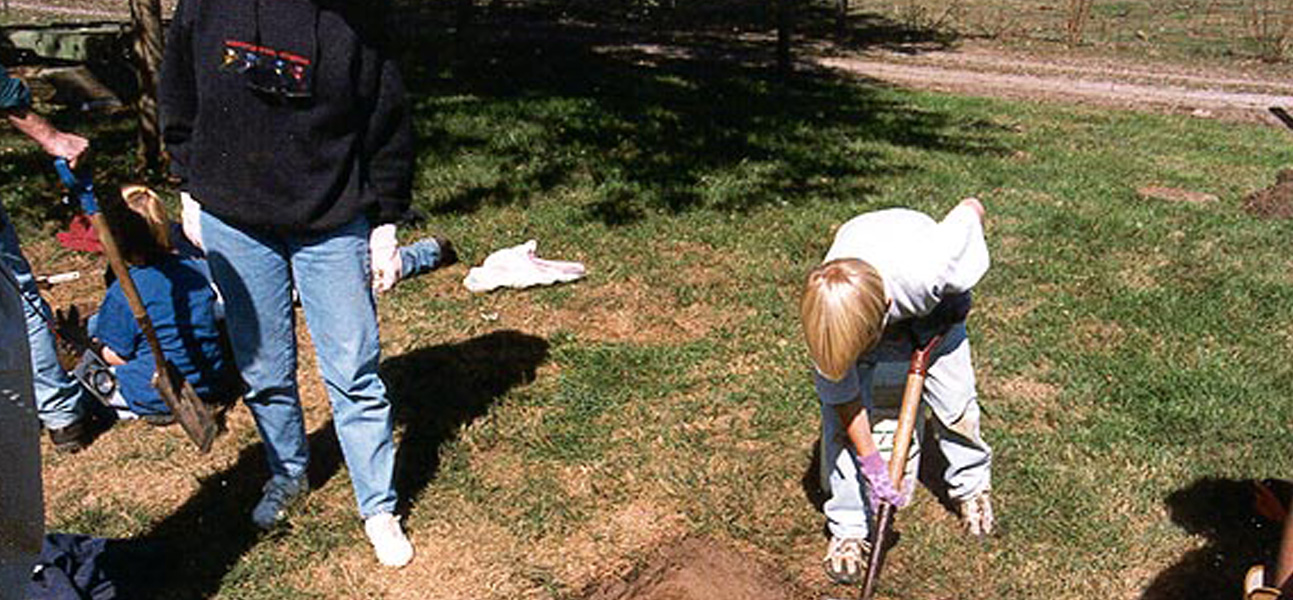 Student bent over, digging hole with shovel, with adult standing to the left.