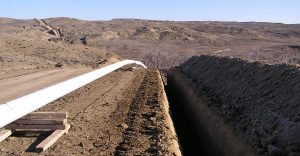 Pipeline trench next to dirt road, with elevated mountainous terrain in background.