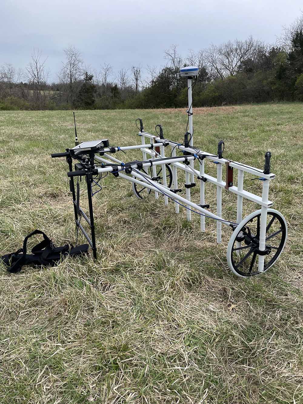 Pushcart magnetometer, a metallic white pole structure resting on two wheels, in open field.
