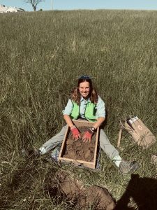 Ashley sitting with soil screen in tall-grass field.