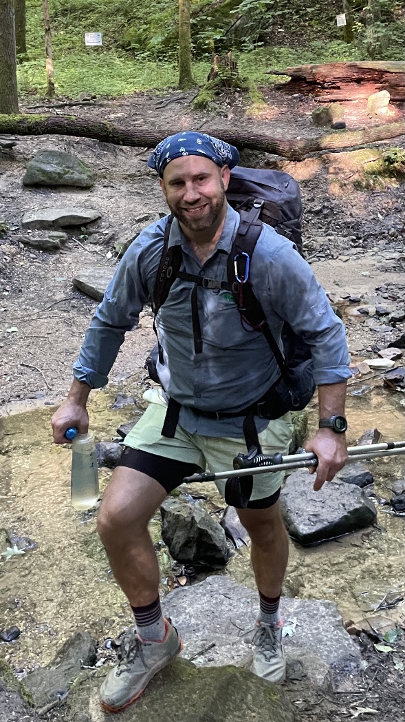 John standing with hiking gear on creek bed.